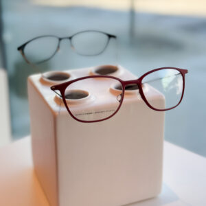 Two eyeglass frames sit atop a decorative vase. Both are rounded, rectangular shapes; one is burgundy and one is black. 